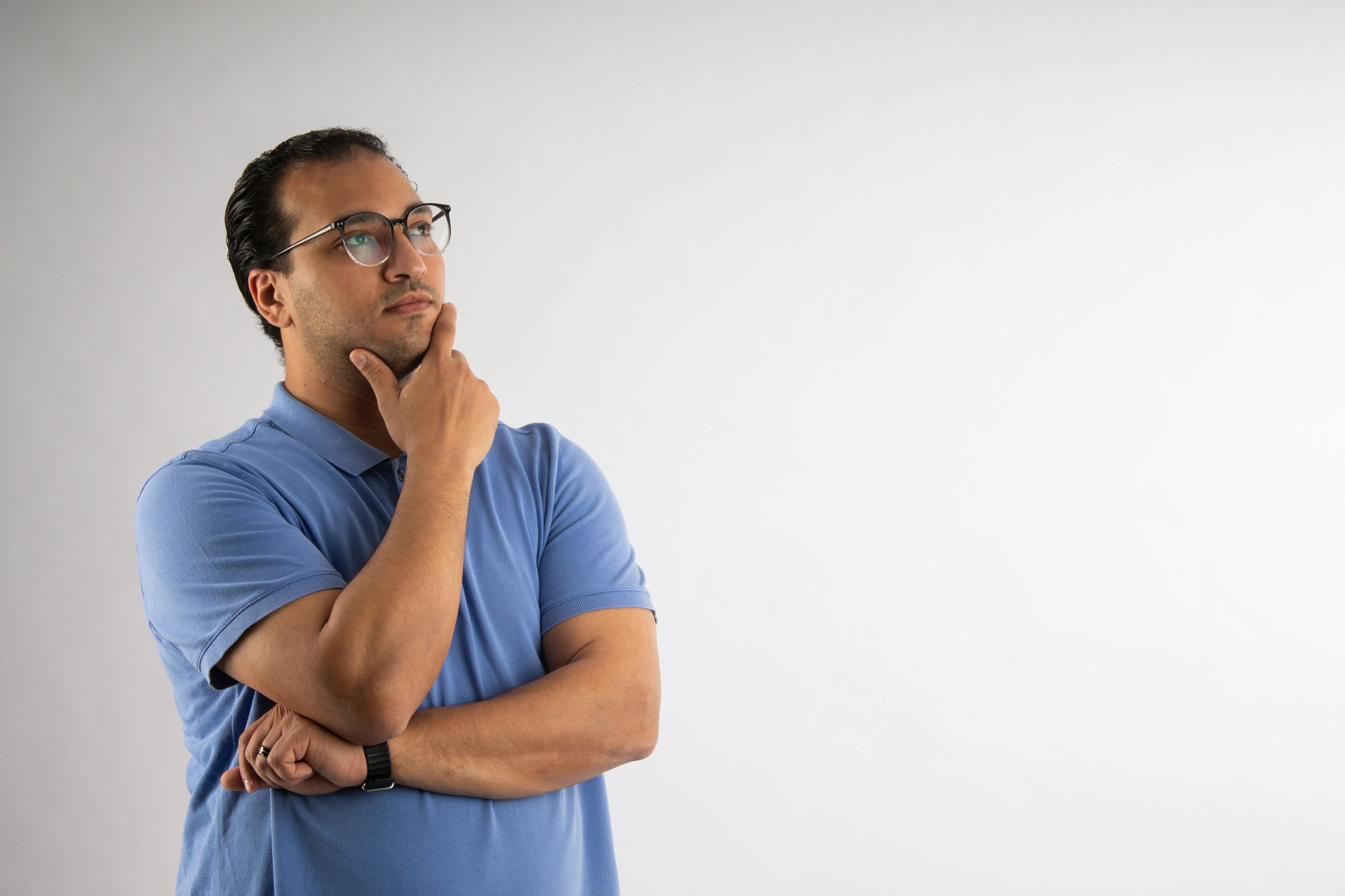 grátis Um homem adulto pensativo, usando óculos e uma camisa polo azul, ponderando sobre um fundo branco. Foto profissional