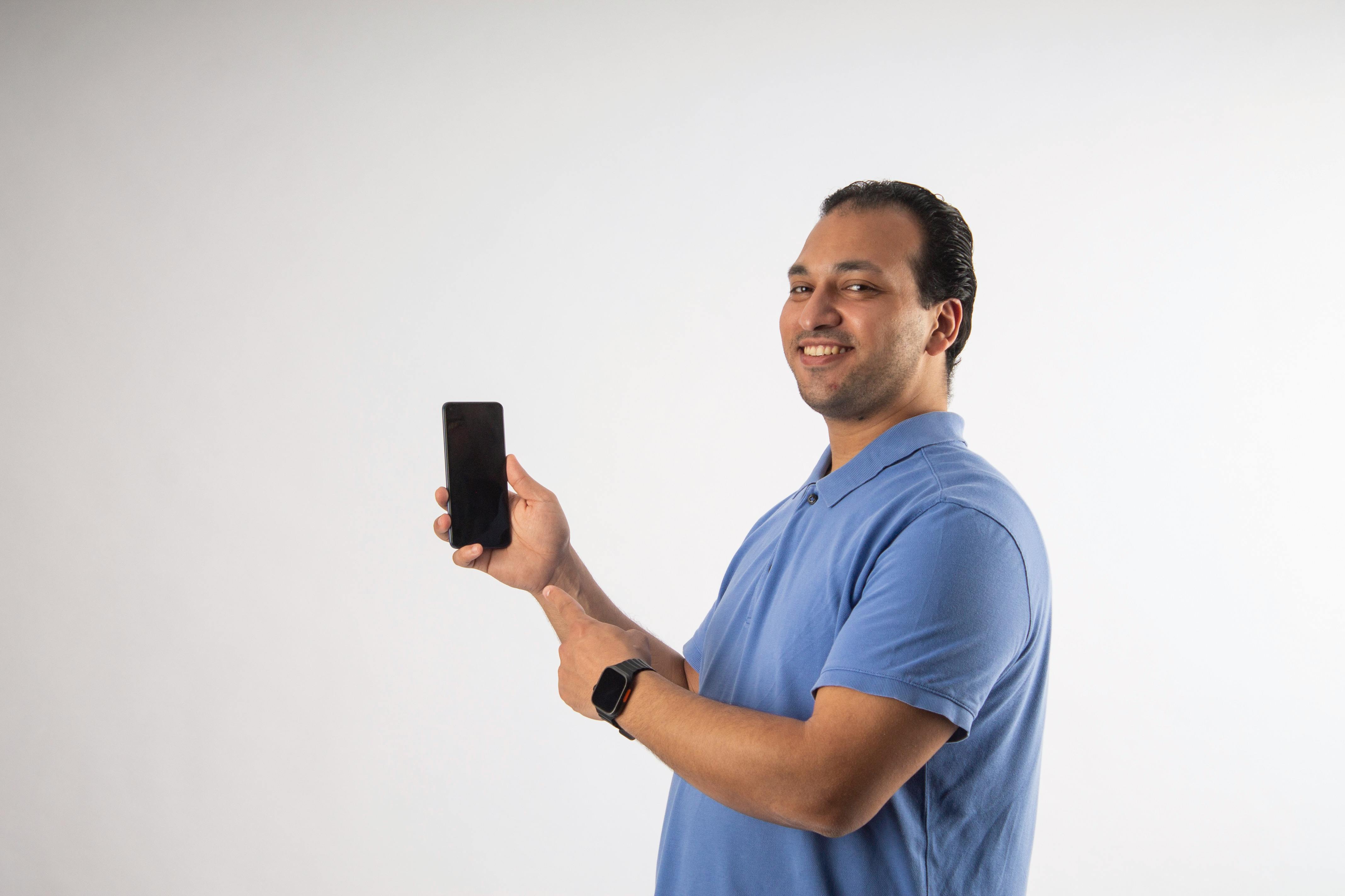 Bezpłatne Uśmiechnięty mężczyzna w niebieskiej koszuli trzyma i wskazuje na czarny smartfon na białym tle. Zdjęcie z galerii