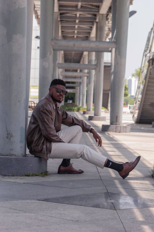 Free Young man in casual attire sitting under a modern bridge, exuding confidence and style. Stock Photo