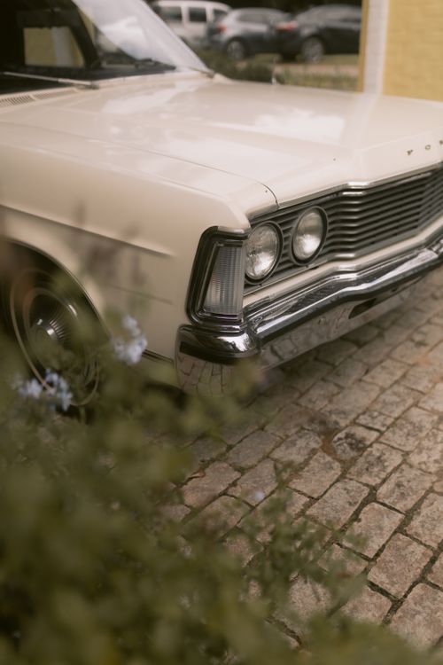 Free Classic vintage white car parked on a cobblestone street with blurred foliage in the foreground. Stock Photo