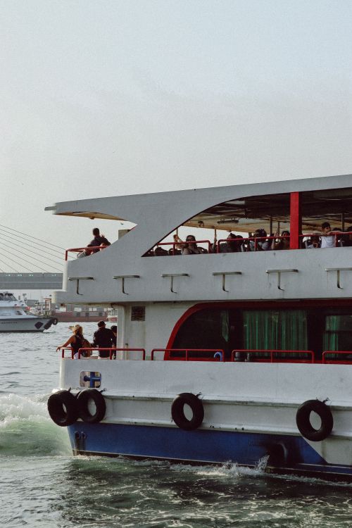 Free A modern ferry with passengers cruising through calm waters near a city's bridge. Stock Photo