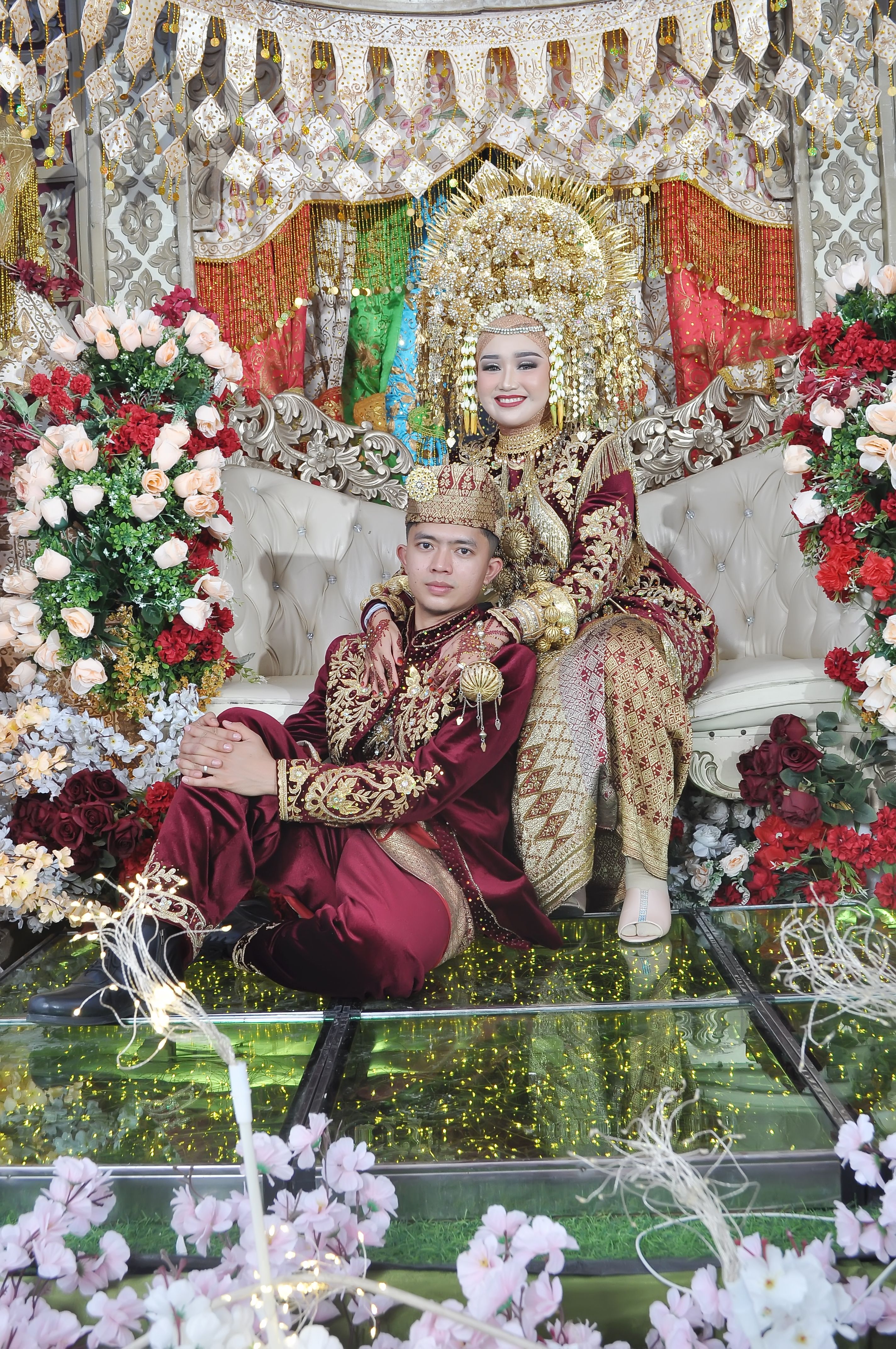 A couple in traditional ornate wedding attire surrounded by vibrant floral decorations.