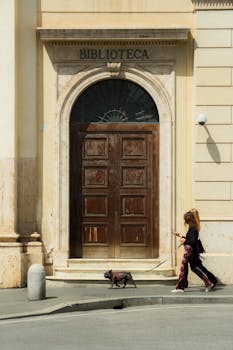 Woman walks dog past historic library entrance on sunny day, capturing urban lifestyle.
