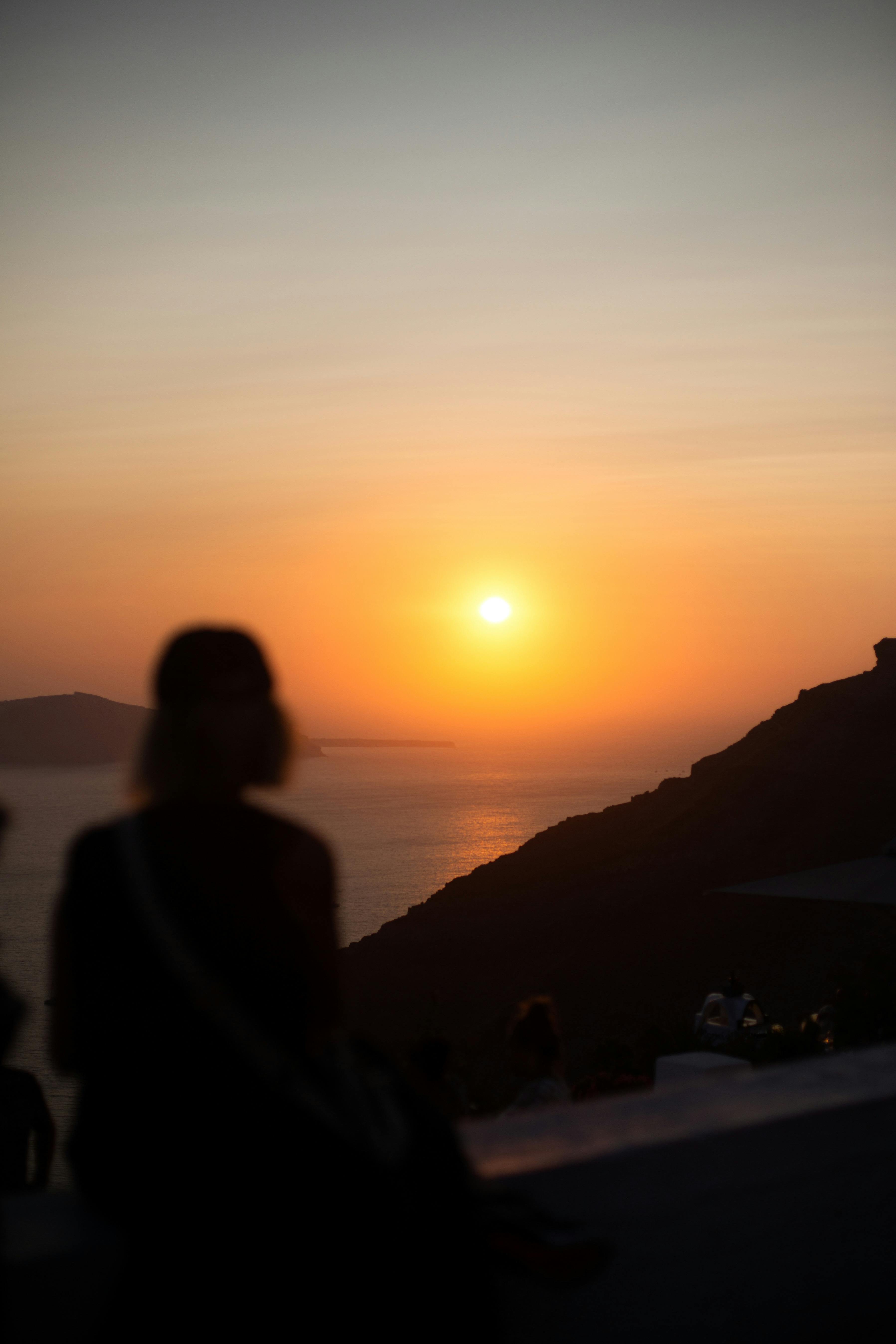 ฟรี ภาพเงาของบุคคลกำลังชมพระอาทิตย์ตกดินที่สวยงามเหนือชายฝั่งของซานโตรินี บรรยากาศสงบและงดงามราวกับภาพวาด คลังภาพถ่าย