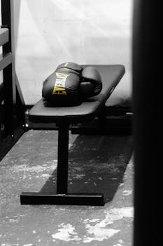Black boxing gloves resting on a gym bench in a monochrome setting, evoking focus and intensity.