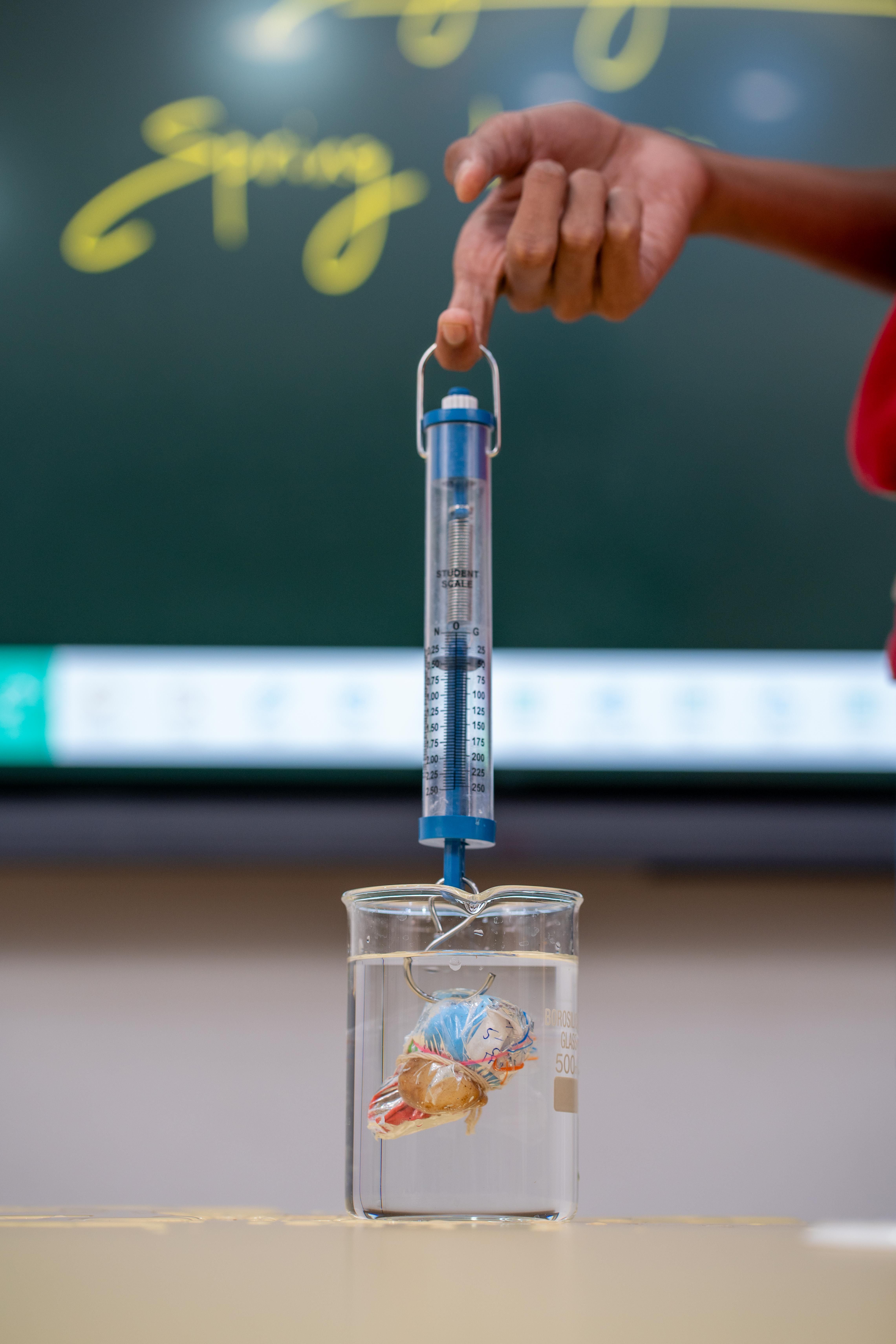 A physics classroom scene showing a hand using a spring with a beaker of water.