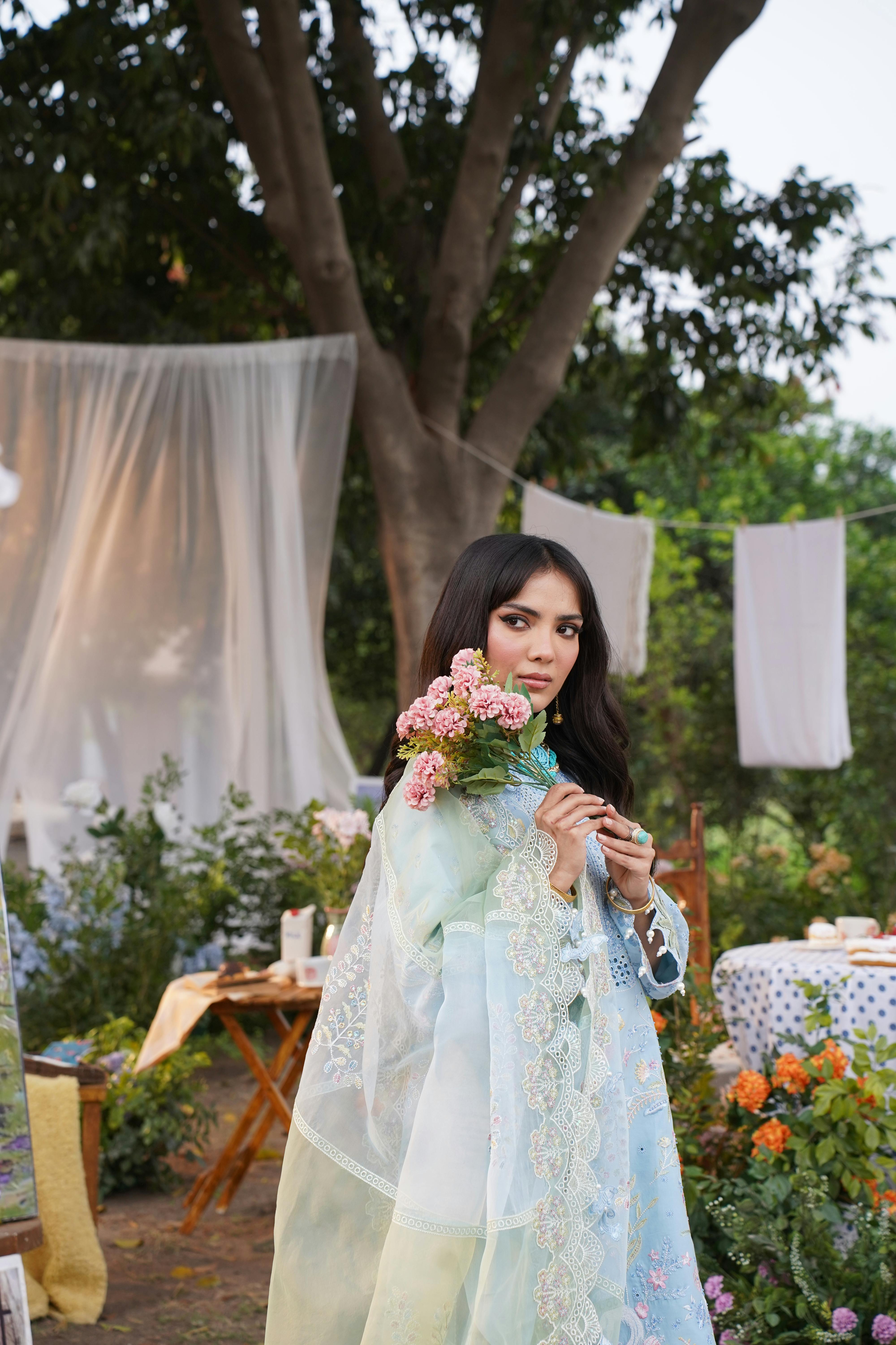 A fashionable woman in a traditional dress holding flowers in an outdoor setting in Lahore, Pakistan.