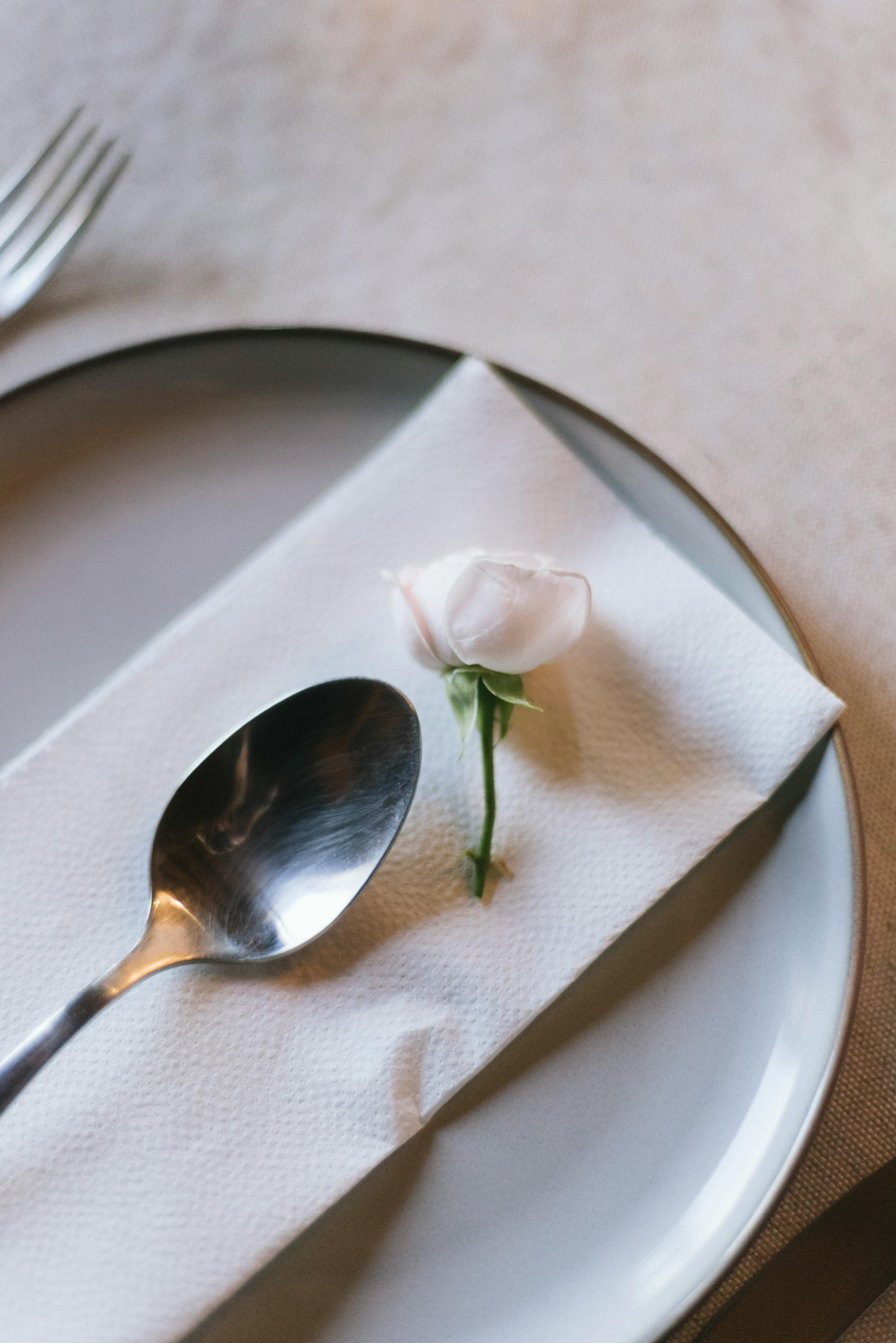 A minimalist table setting with a plate, napkin, spoon, and delicate rosebud.