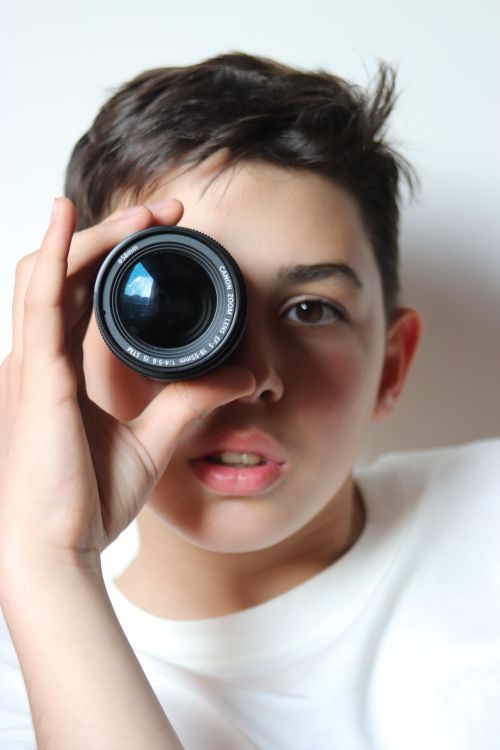 Free Teen boy peering through a camera lens with curiosity and focus. Stock Photo
