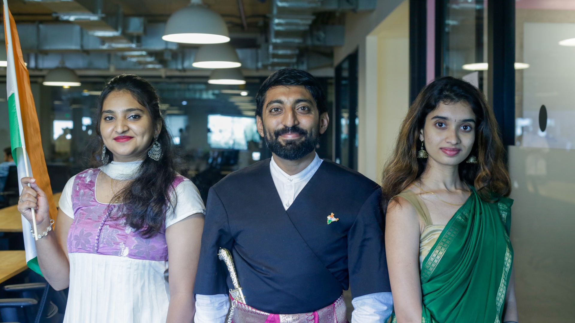 Three adults in traditional Indian clothing posing in an office setting with the Indian flag.