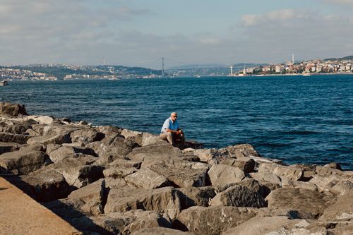 Free A man enjoys a quiet moment by the rocky shores overlooking Istanbul and the Bosphorus Bridge. Stock Photo