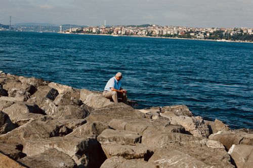 Free An elderly man sits on rocks by the Bosphorus, enjoying a peaceful moment with a cityscape backdrop. Stock Photo