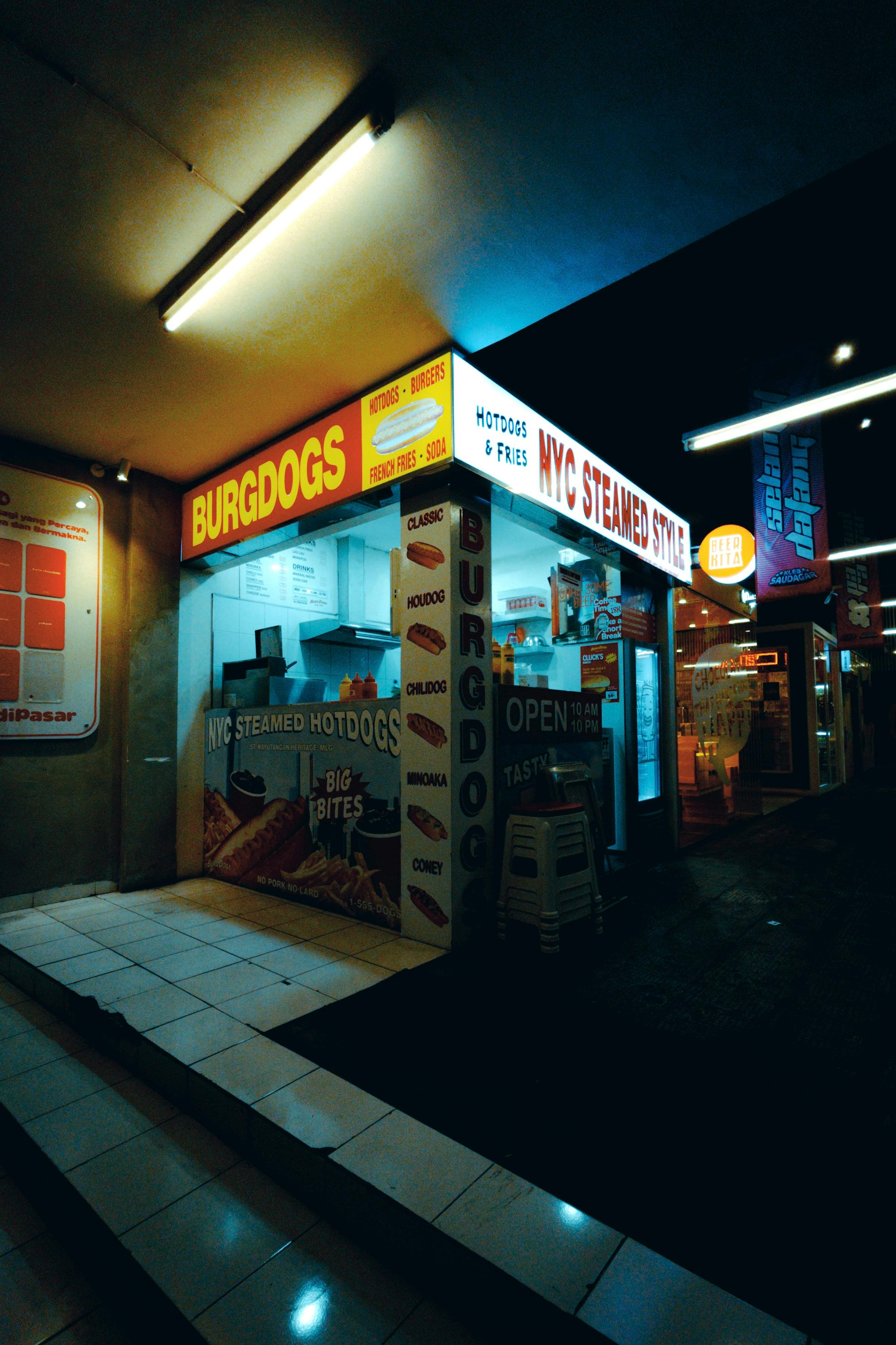 grátis Uma barraca iluminada de cachorro-quente cozido no vapor em Nova York servindo comida de rua sob luzes de neon durante a noite. Foto profissional