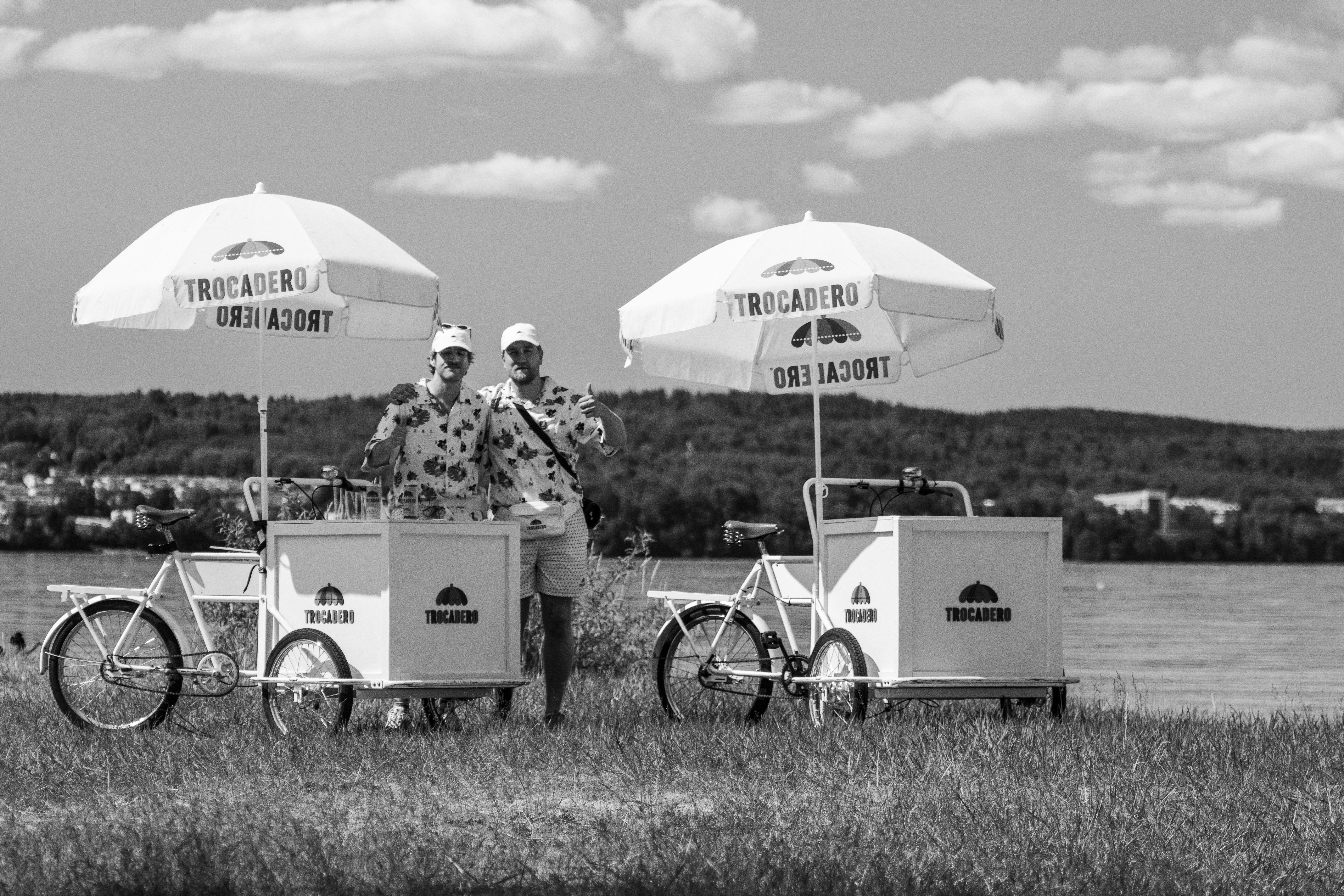 Two ice cream vendors stand with bicycles near a lake under umbrellas labeled 'Trocadero'.