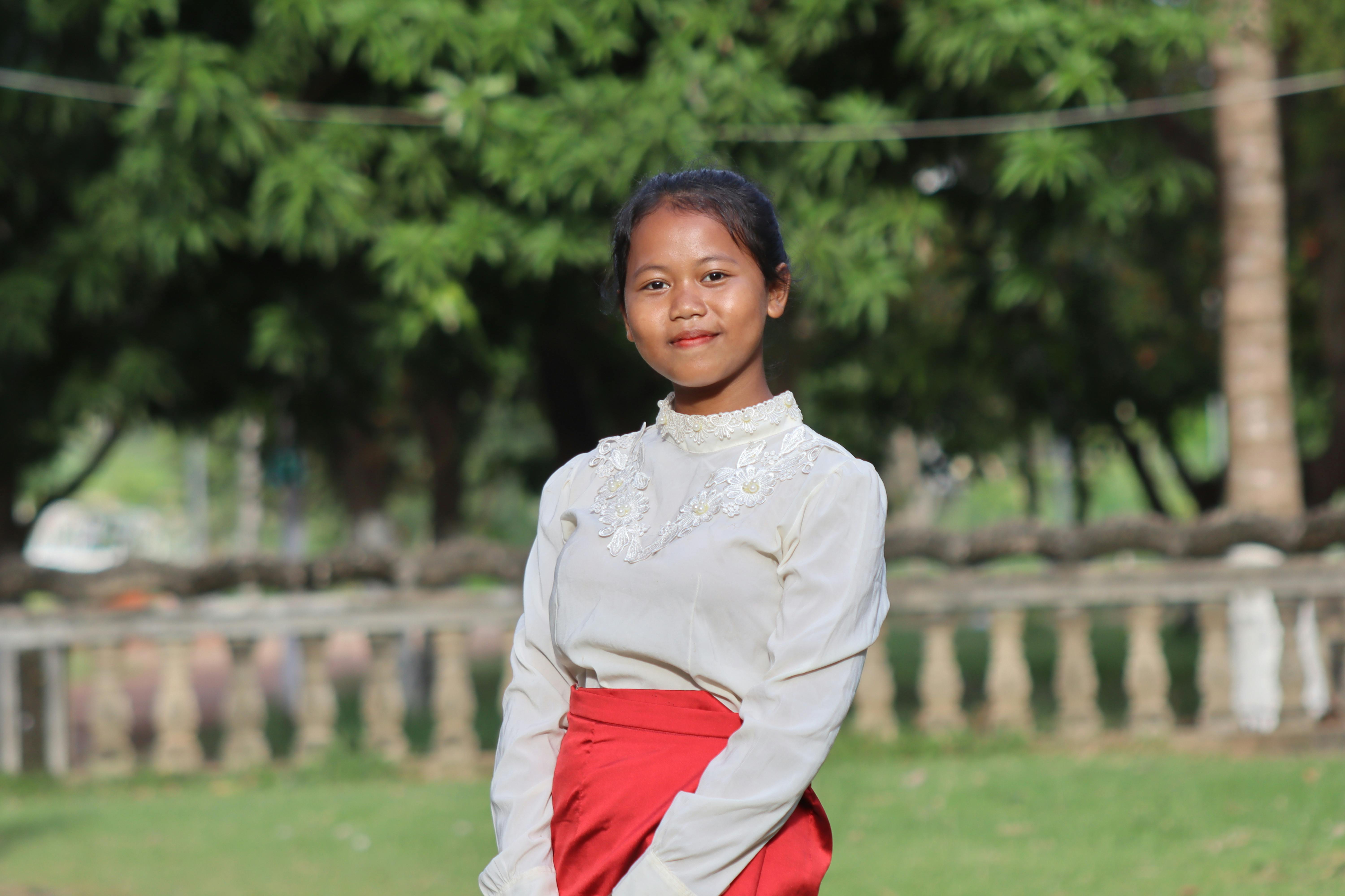 A young Cambodian woman wearing traditional attire poses outdoors with a bright smile, surrounded by lush greenery.
