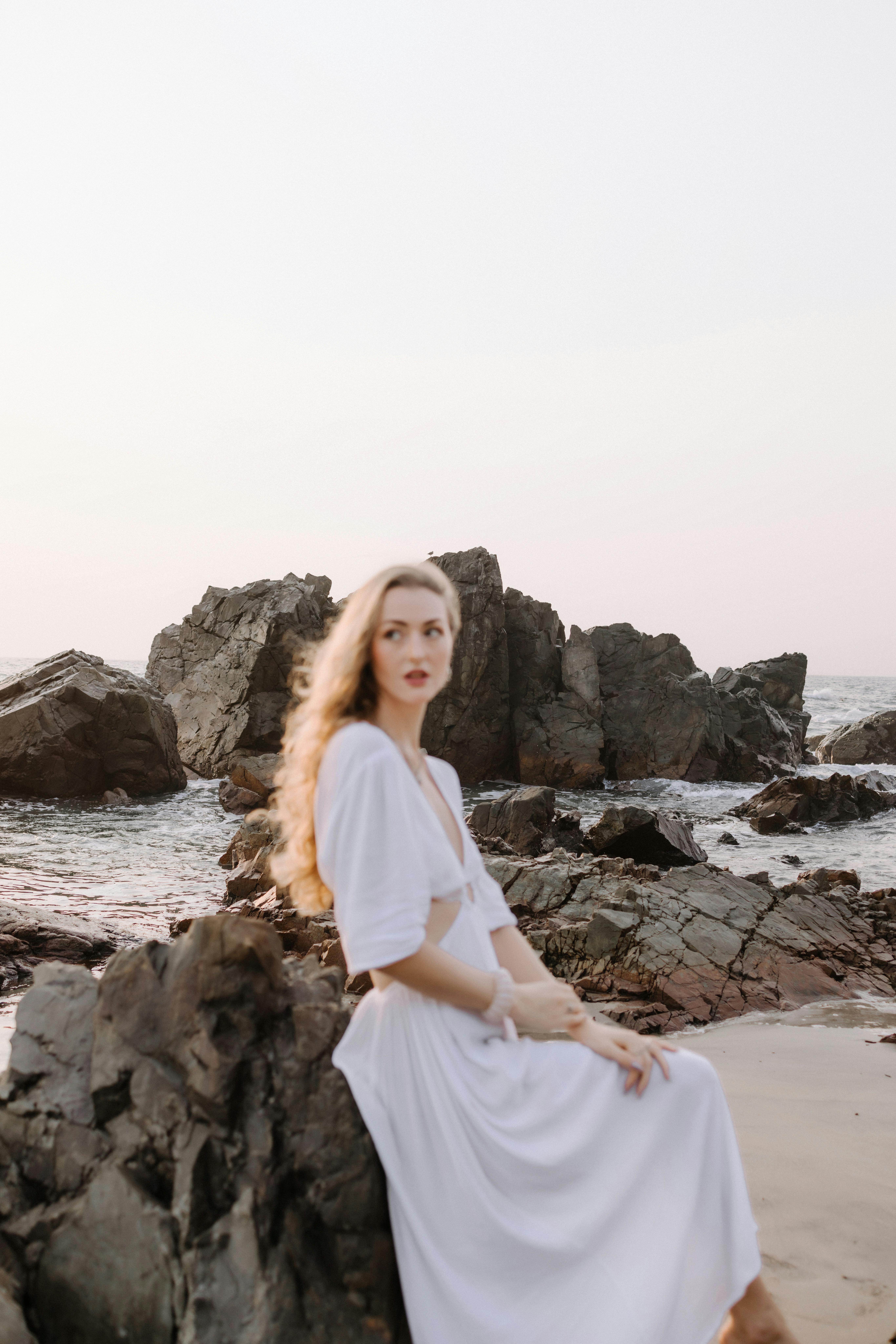 穿着白色连衣裙的优雅女子坐在宁静海滩的岩石旁