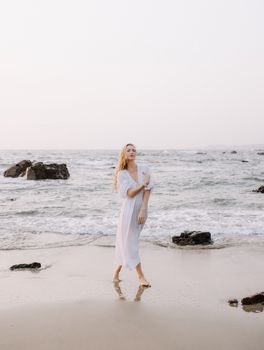 A woman in a white dress strolls gracefully along a serene beach at sunset.