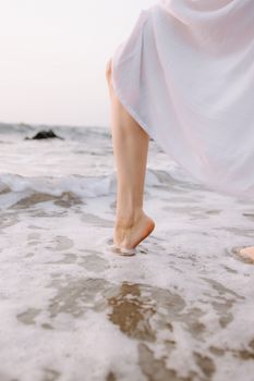 A woman in a white dress walks barefoot along the sandy ocean shore, calming and serene.