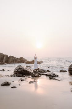 Person meditating on rocky beach during sunset, creating a calm and serene atmosphere.
