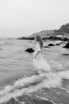 Artistic black and white photo of a woman in a flowing dress walking along the beach.