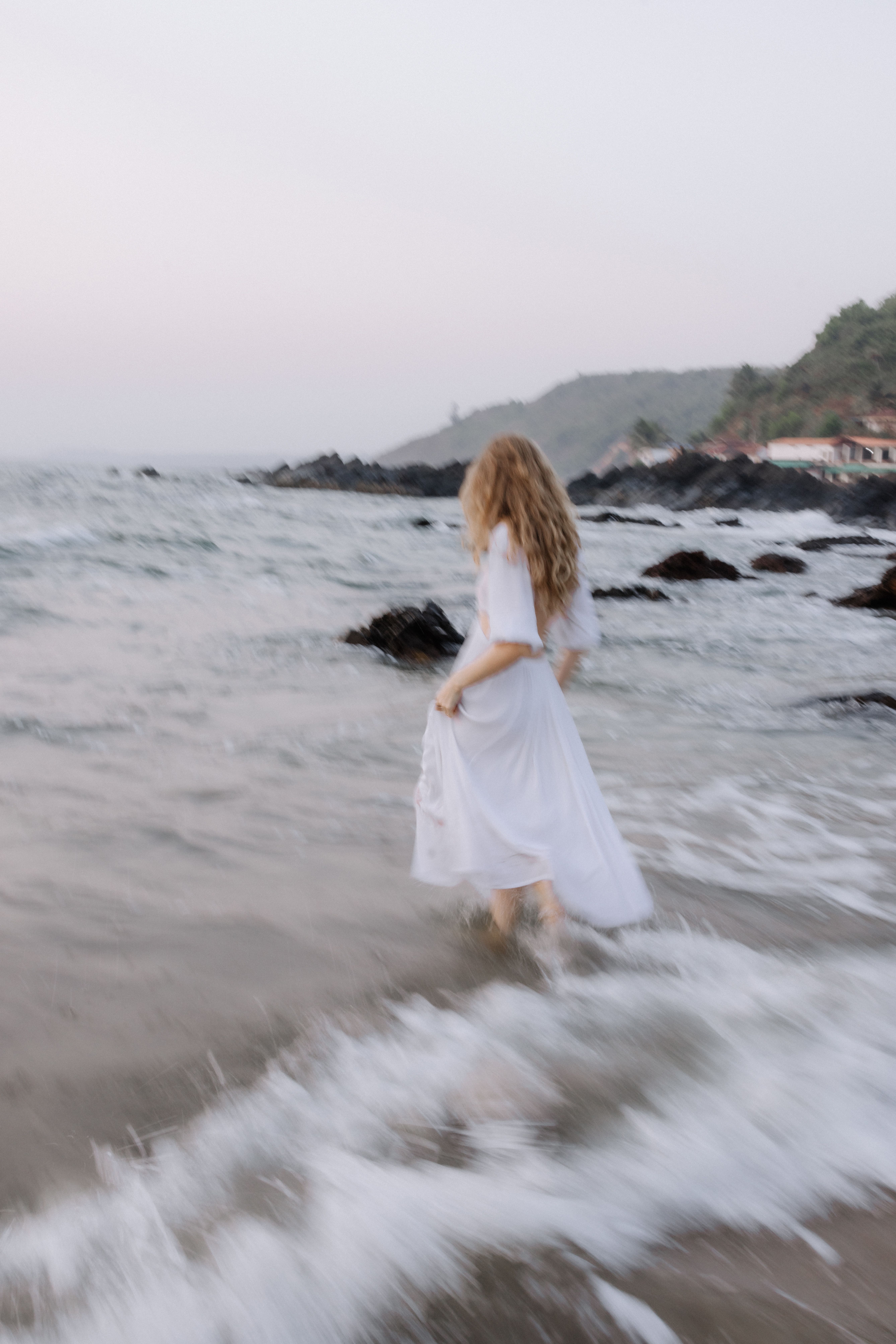 Blurry image of a woman in white dress walking along the ocean shore at twilight.