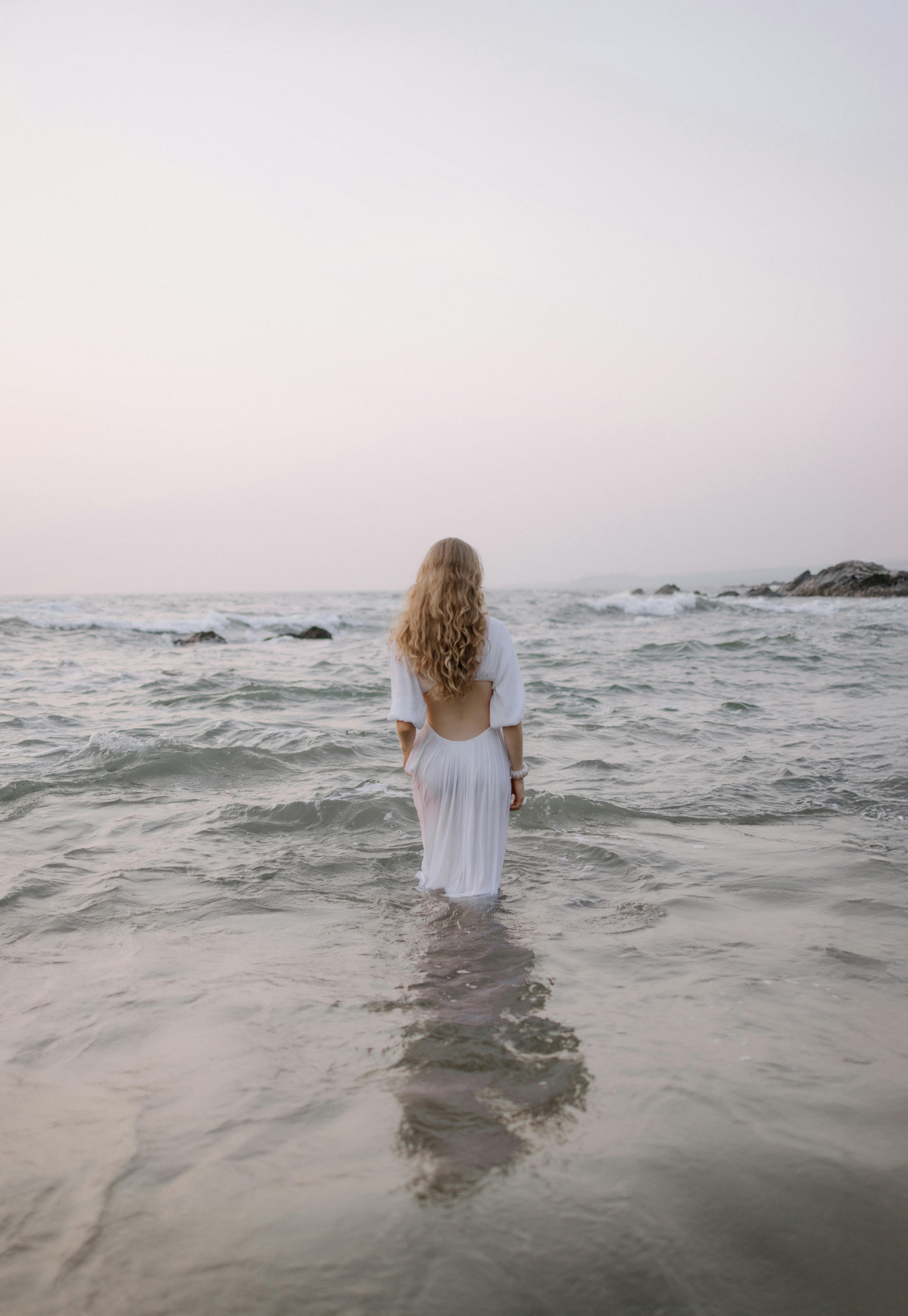 A woman in a white gown stands in a serene ocean under a pastel sunset sky.