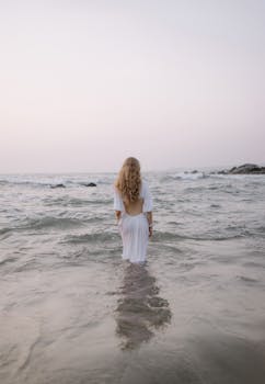A woman in a white gown stands in a serene ocean under a pastel sunset sky.