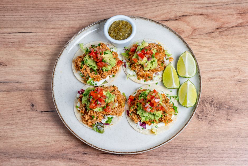 Shredded chicken taco filling piled onto corn tortillas and topped with cilantro, diced onion, and lime wedges, with the slow cooker visible in the background.