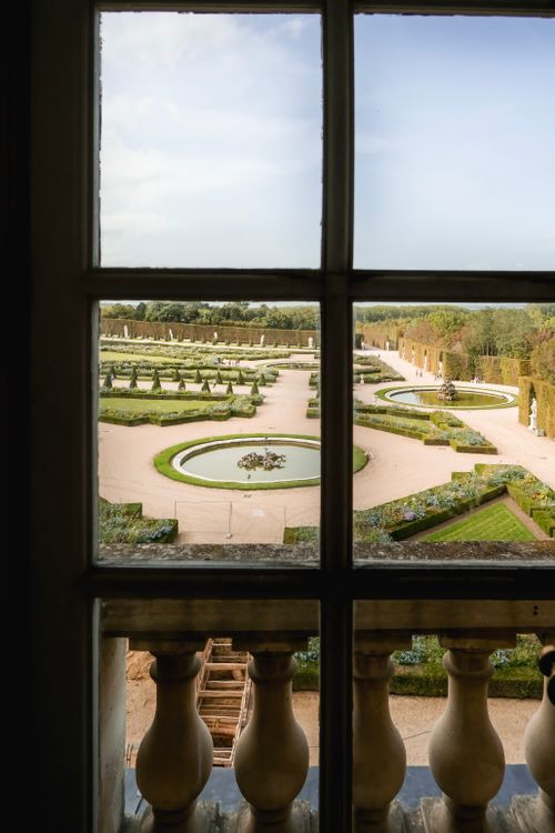 Free A picturesque view of Versailles gardens seen through a window, capturing the elegance of French landscape design. Stock Photo