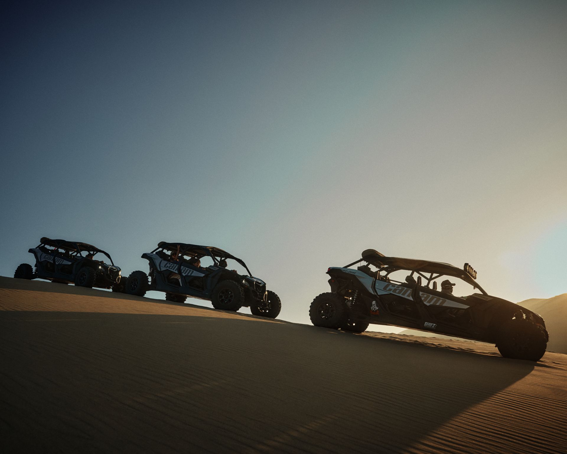 Three off-road vehicles traverse sand dunes against a striking sunset.