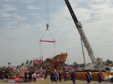 A colorful traditional boat being lifted by a crane during a cultural ceremony outdoors.