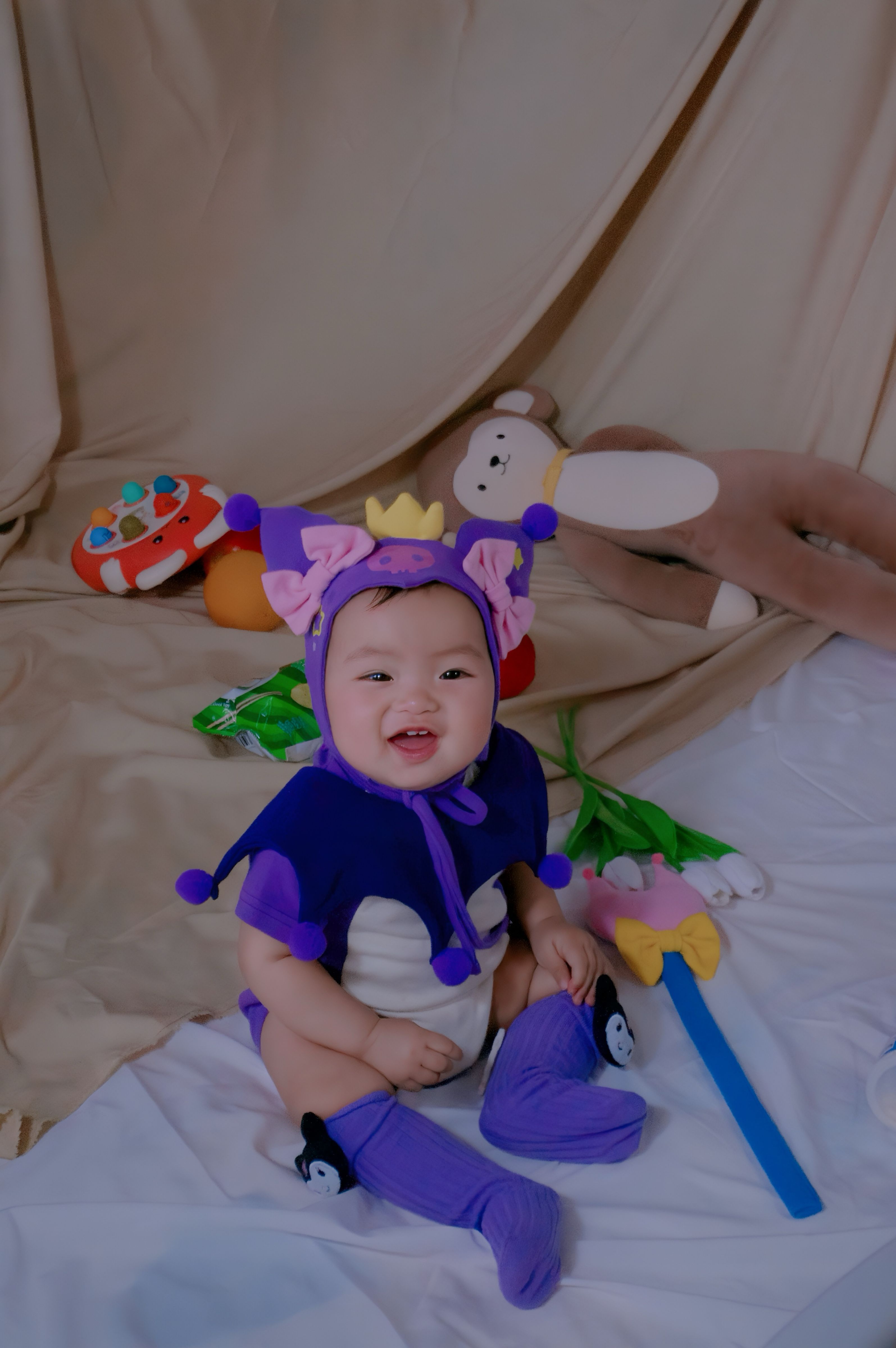 Cute baby in a vibrant jester outfit surrounded by toys, indoors setting.