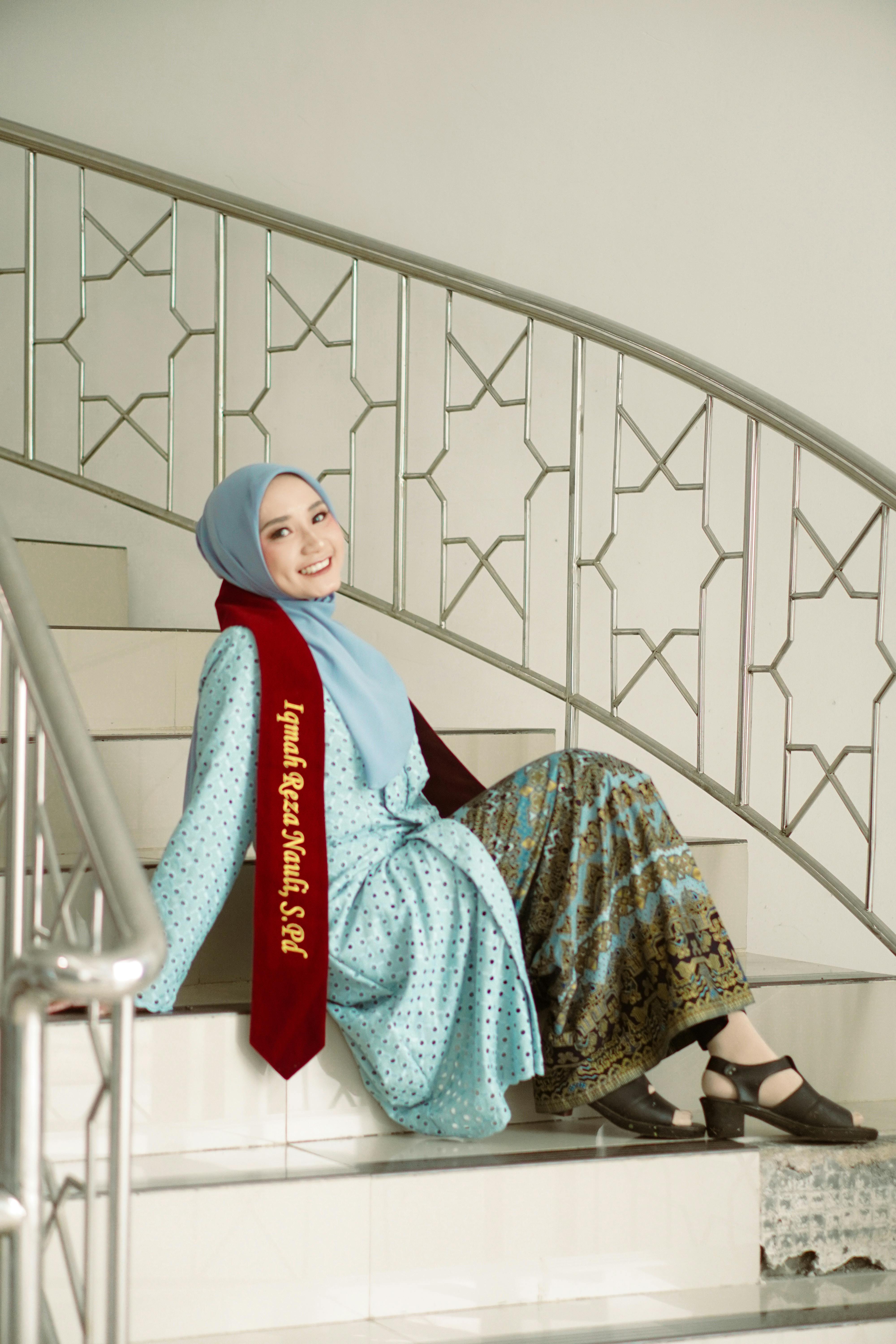 A young woman in traditional dress with a hijab, smiling on a staircase, celebrating her graduation.