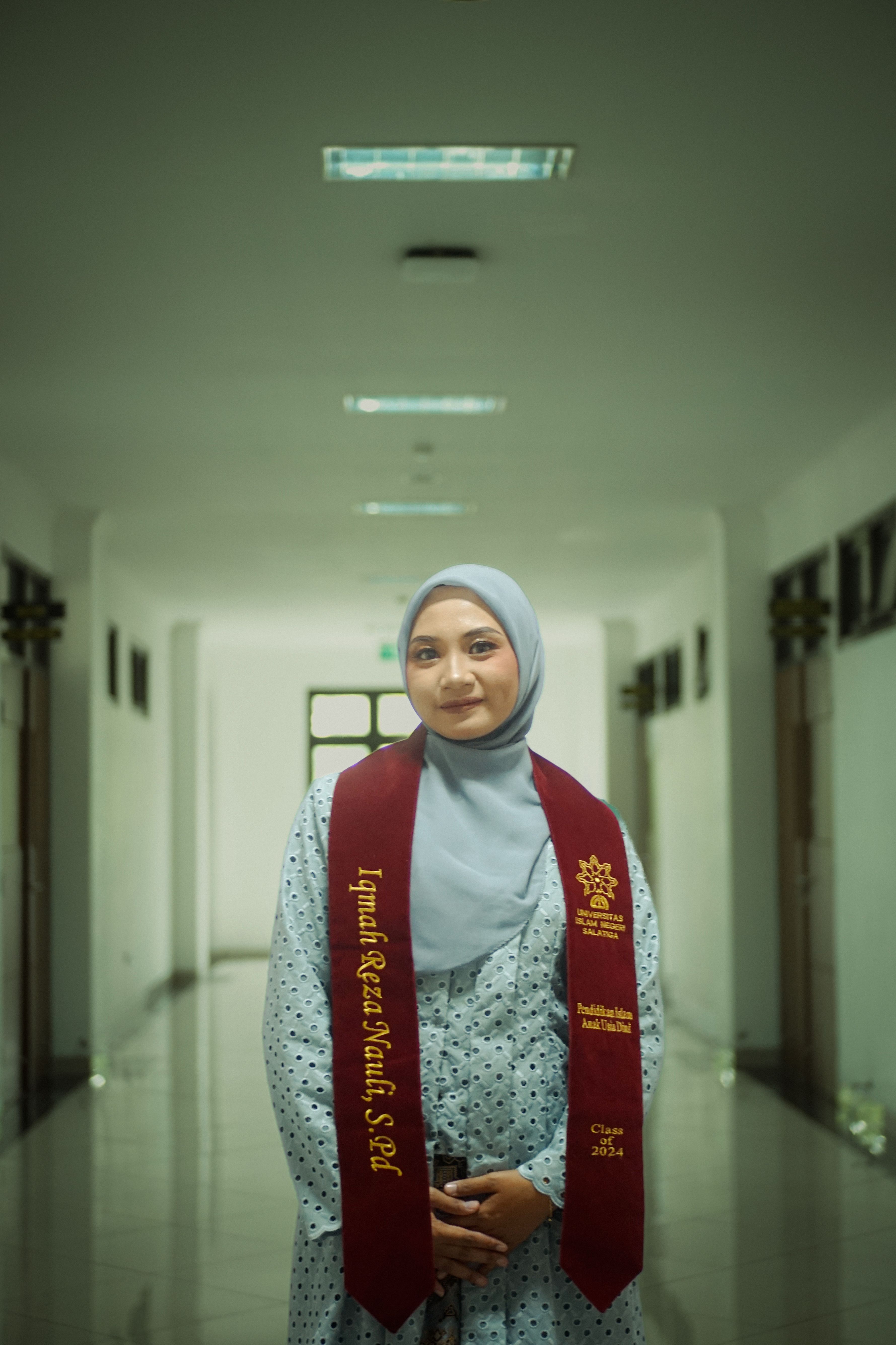 Portrait of a young woman wearing a hijab and graduation stole, smiling in a hallway.