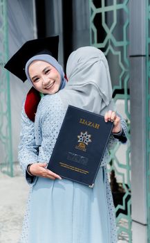 Two women celebrating graduation with a joyful hug, wearing hijabs and holding diploma. - Fotografer Sidoarjo