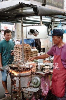 Street vendor preparing char kway teow with sparks flying from a wok in an outdoor market.