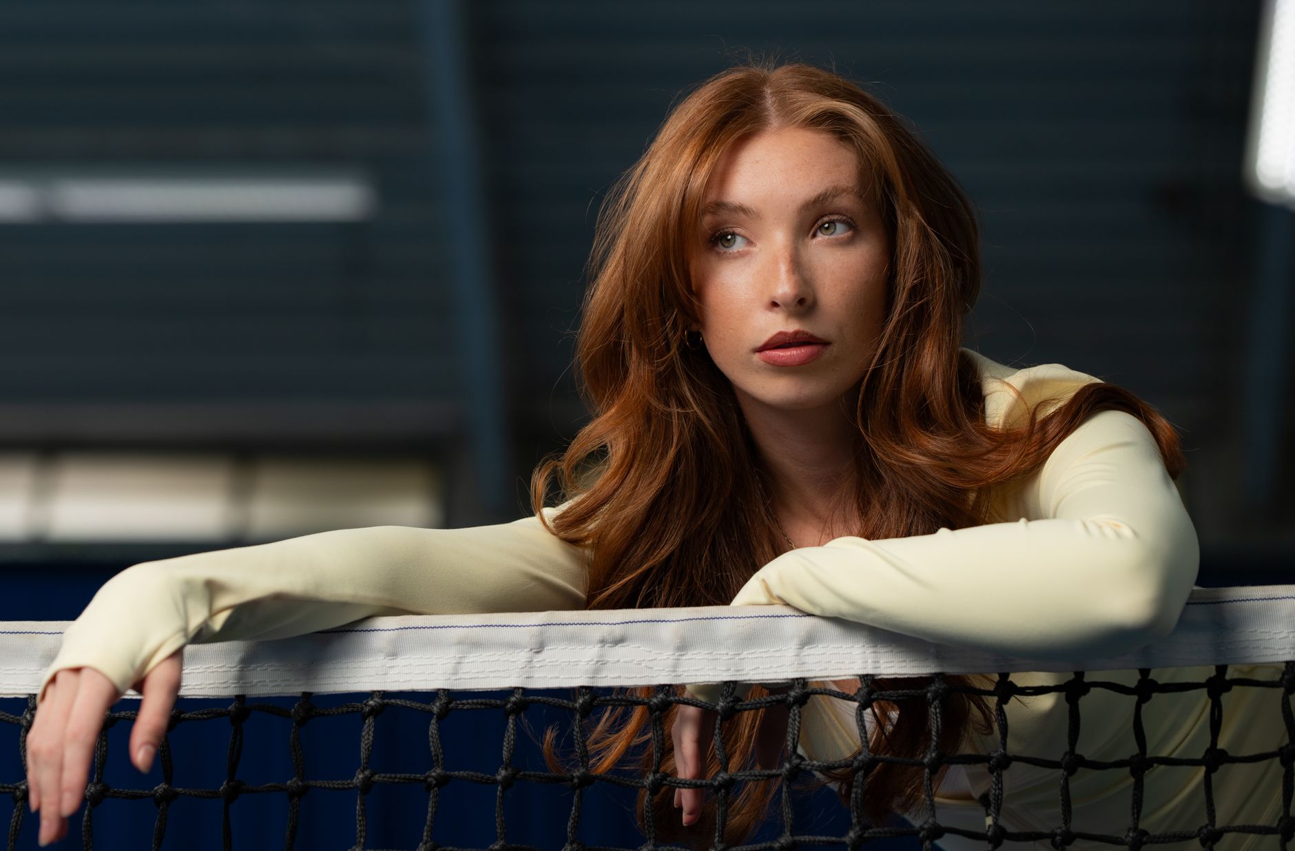Portrait of a young woman leaning on a tennis net indoors. Natural pose and thoughtful expression.