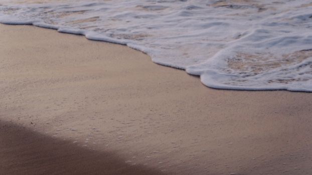Soft waves gently lap the sandy shore under the warm glow of sunset, creating a peaceful scene.