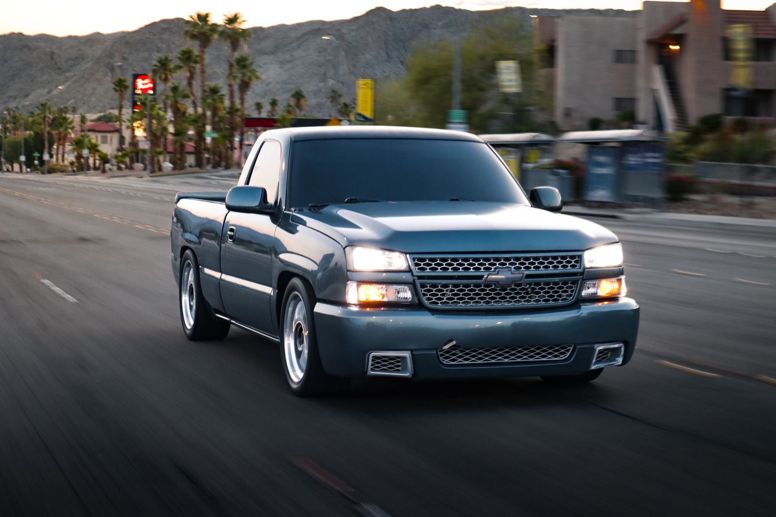 A blue pickup truck speeds down a desert highway lined with palm trees.