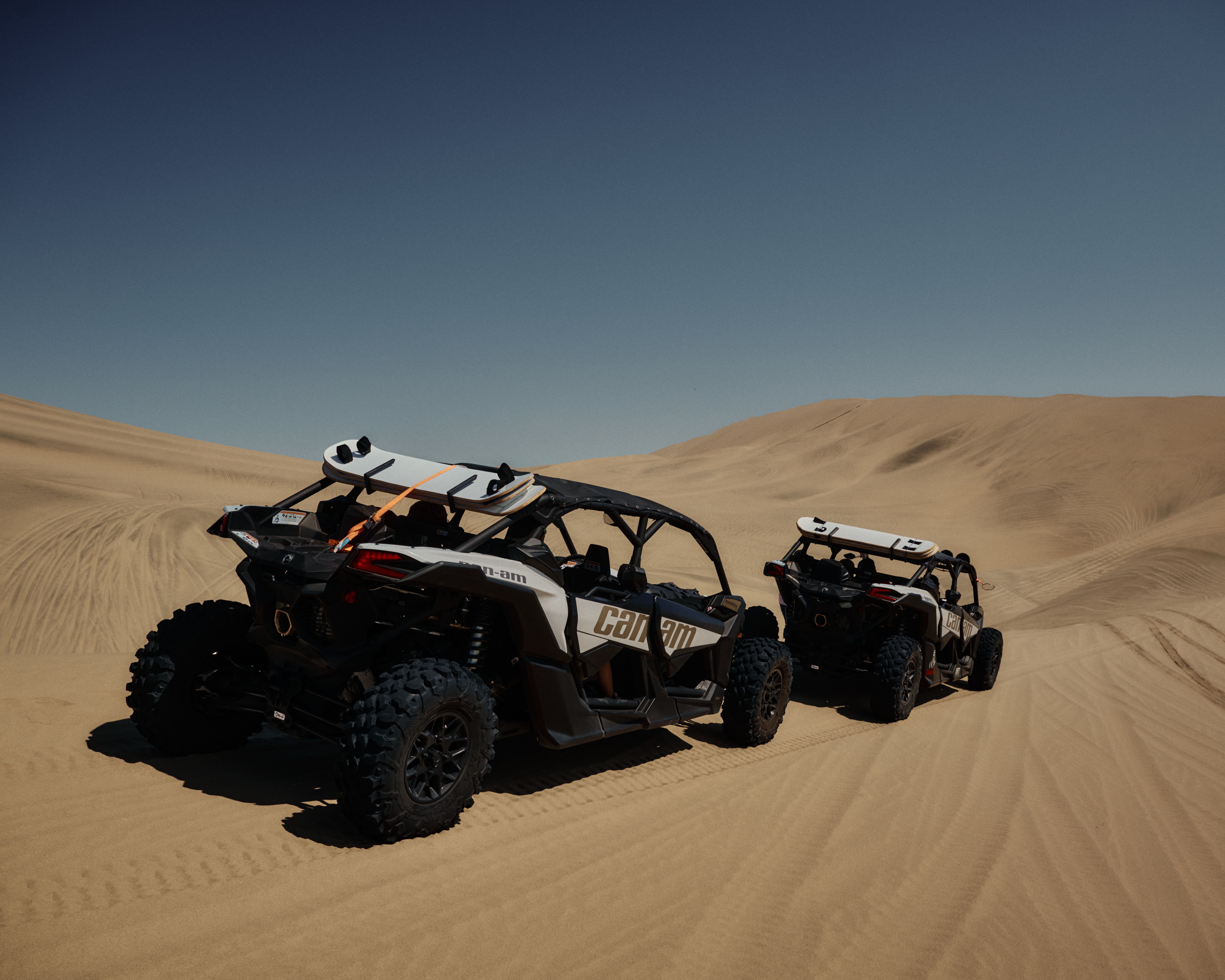 Two off-road vehicles navigate the vast desert sand dunes under a clear blue sky.