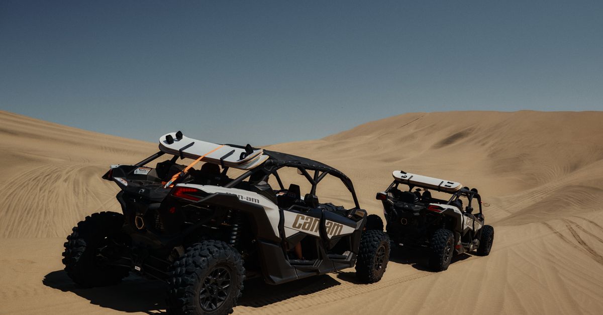 Two off-road vehicles navigate the vast desert sand dunes under a clear blue sky.