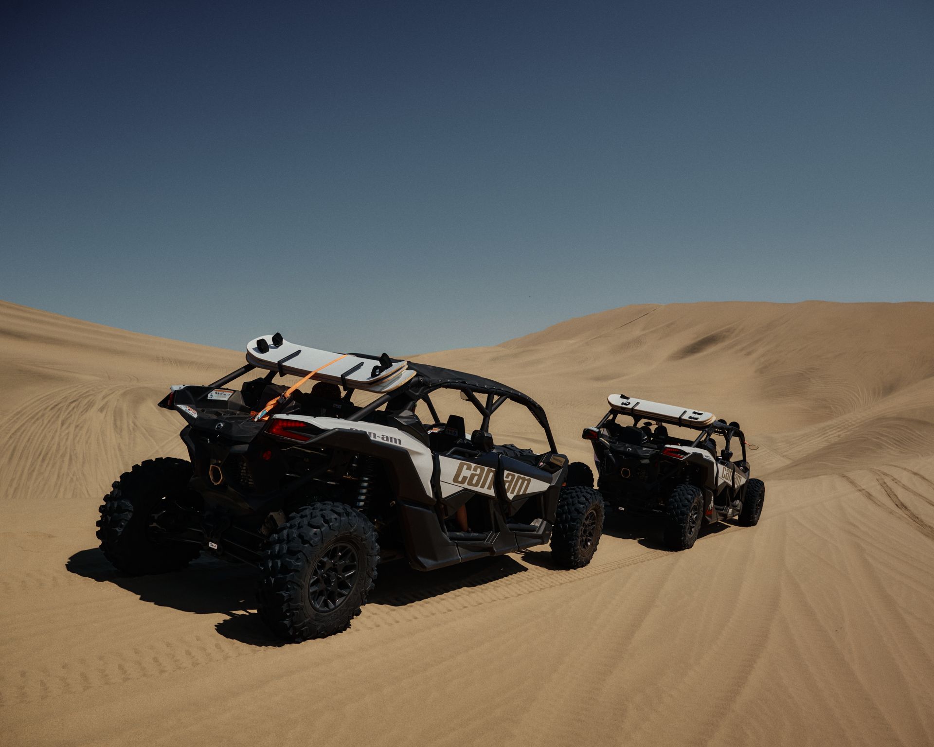 Two off-road vehicles navigate the vast desert sand dunes under a clear blue sky.
