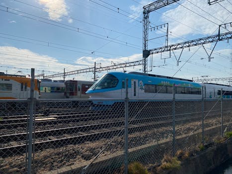 A modern blue train traveling through a rail yard under a sunny sky with overhead power lines.