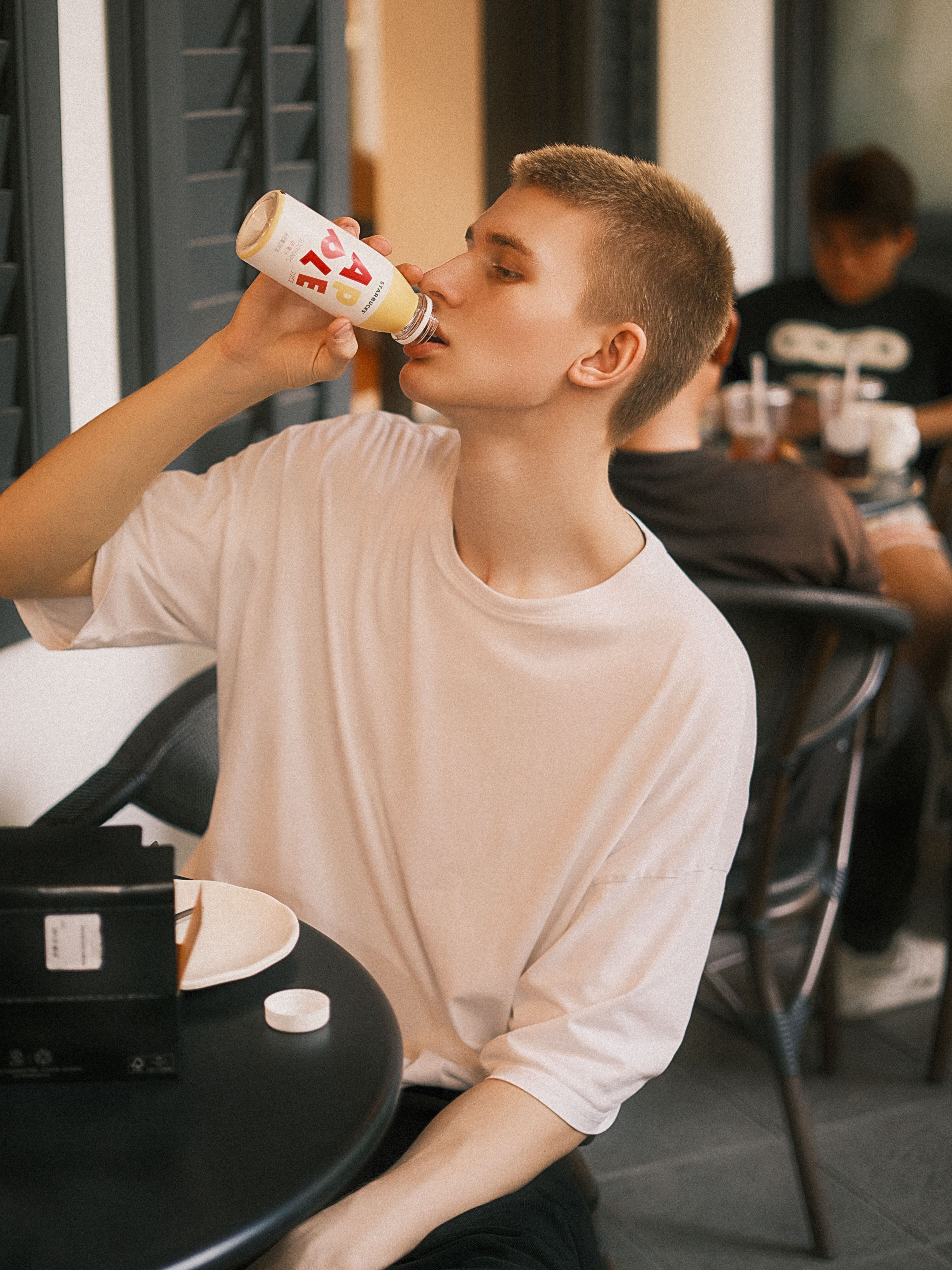 A young man drinks from a can while seated at a casual cafe with friends, creating a relaxed and social atmosphere.