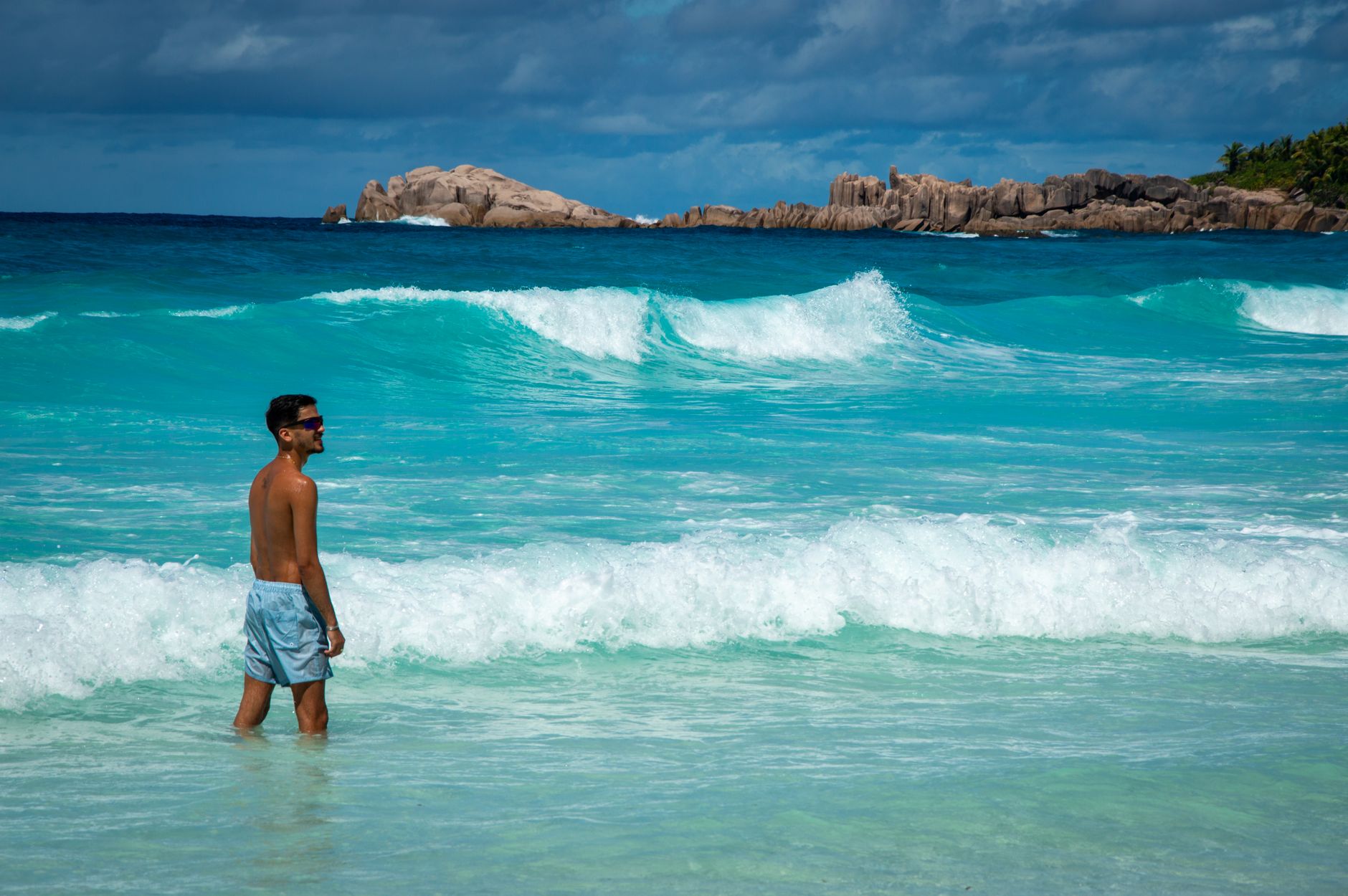 Man standing in the turquoise waters of Anse Source d'Argent, Seychelles, under clear summer skies.