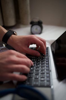 Close-up of hands typing on a laptop keyboard, clock in the background.