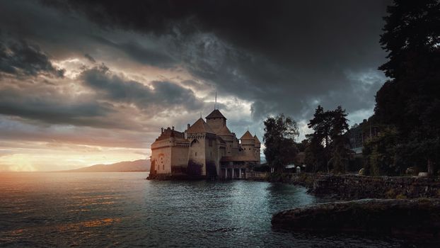 Dramatic view of Chillon Castle on Lake Geneva during sunset with moody clouds.