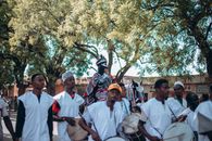 A vibrant African parade featuring drummers and a horseman in traditional attire under lush trees.