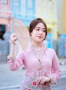 Young woman holding fan, wearing traditional pink Asian attire in colorful street setting. Calm and elegant expression.