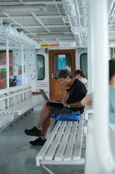 Man sitting on a ferry bench using a laptop, casual travel setting.